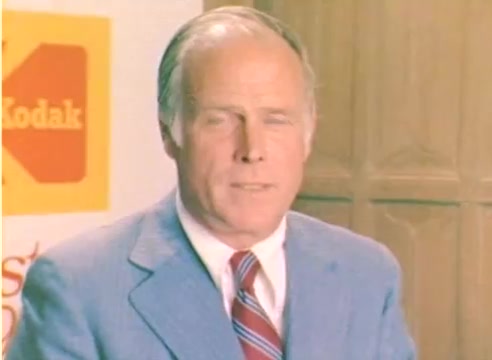 The still features a man in a light blue suit with a tie, standing against a backdrop with Kodak branding. The setting suggests a promotional context related to Kodak's products for the 1977 Christmas season. The color negative reflects the vibrant style typical of the late 1970s. The man appears to be delivering a message, likely promoting Kodak's offerings during the holiday season.