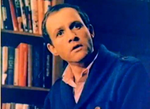 The still from the 16mm silent color negative features a man with a surprised or contemplative expression, dressed in a blue sweater over a pink collared shirt. He is positioned against a background of bookshelves filled with various books, suggesting a setting that might be conversational or reflective. The film is part of a Kodak sales project leading up to the 1977 Christmas season, showcasing the talents of both Michael Landon and Dick Van Dyke.