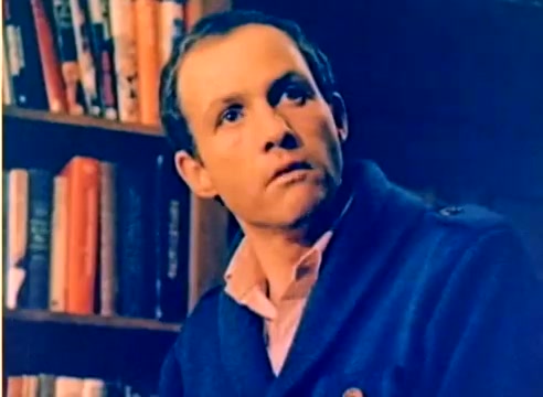The still features a man with a thoughtful expression, wearing a blue sweater over a light-colored shirt, seated in front of a bookshelf filled with books. The color is vibrant, characteristic of a silent 16mm negative from a Kodak sales film. The context suggests it is part of promotional content leading up to the 1977 Christmas season, showcasing the personalities of Michael Landon and Dick Van Dyke.