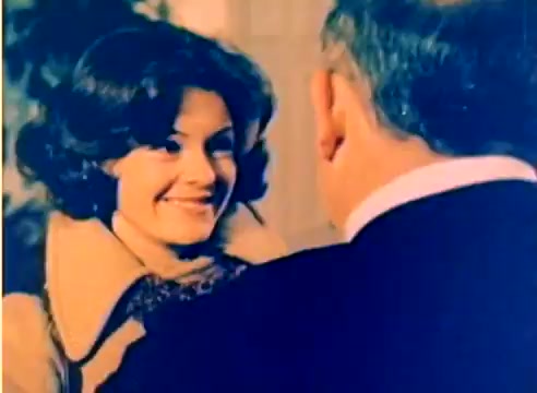 The still from the 'Kodak 1977 Sales Film' features a woman with a warm smile, dressed in a stylish outfit, interacting with an older man whose back is visible. The setting appears festive, likely capturing the holiday spirit leading up to Christmas. The colors are bright, typical of the vibrant Kodak film style of the era. The scene conveys a sense of joy and connection, fitting for a promotional film.