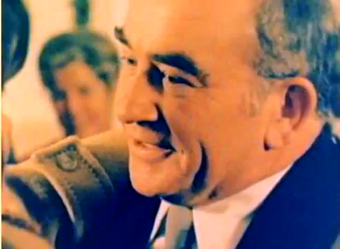 The still features a close-up of a smiling man, exuding warmth and friendliness. He appears to be in a celebratory setting, likely during a holiday gathering. The colors are vibrant, typical of a Kodak film from that era. In the background, blurred figures suggest a festive atmosphere, contributing to the overall cheerful tone of the scene.