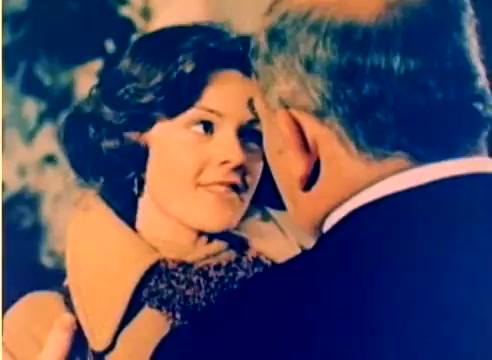 The still from the 'Kodak 1977 Sales Film' captures a close-up moment between two characters, likely a romantic or sentimental exchange given the expressions. One character, a woman with curly hair, appears joyful and engaged, while the other, a man whose face is partially obscured, leans in, suggesting intimacy. The colors are vibrant, typical of Kodak's film quality, and the soft focus enhances the warm, nostalgic feel of the scene, emphasizing the holiday theme.