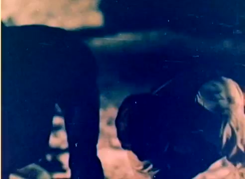 The still captures a moment from a 16mm silent color negative featuring two figures, likely Michael Landon and Dick Van Dyke. The setting appears to be outdoor, possibly during a winter scene, as implied by the context of a Kodak sales film leading up to the 1977 Christmas season. The figures are engaged in an activity close to the ground, but the silent nature of the film and the low-contrast color tone render their actions and expressions ambiguous.