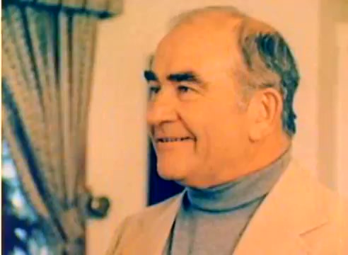 The still depicts a smiling man with a friendly demeanor, casually dressed in a light-colored suit and a turtleneck. The setting appears warm and inviting, indicative of a festive atmosphere. This image is part of a silent color negative from a Kodak sales film, emphasizing the promotional aspects leading into the 1977 Christmas season.