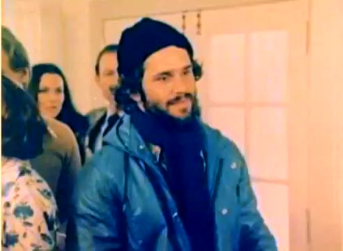 The still from the 1977 Kodak sales film features a man with curly hair wearing a blue jacket, a black scarf, and a beanie, standing among a group of people. The scene conveys a warm, festive atmosphere, likely capturing a moment leading up to the Christmas season. The film's color palette, typical for the era, adds a nostalgic touch. The individuals around him appear engaged in conversation, enhancing the social setting of the scene.