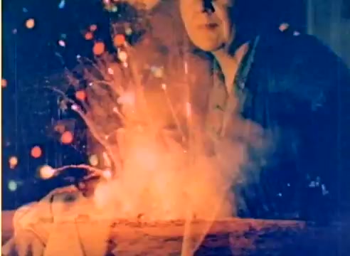 The still captures a festive scene from the 1977 Kodak sales film, featuring a warm glow from a fire or fireworks. There are colorful, blurred lights in the background, suggesting a Christmas ambiance. The focus is on a male figure who appears engaged with the glowing scene, likely portraying a moment of joy or celebration. The colors are vibrant, reflecting the holiday spirit.