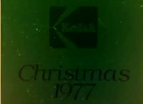 The still from the Kodak 1977 Sales Film features the iconic Kodak logo and the text 'Christmas 1977.' It suggests a promotional film aimed at showcasing Kodak products during the holiday season, associated with notable figures Michael Landon and Dick Van Dyke. The image is captured from a 16mm silent color negative, likely emphasizing the warmth and nostalgia of the era. The background has a green tint, characteristic of older film stock.