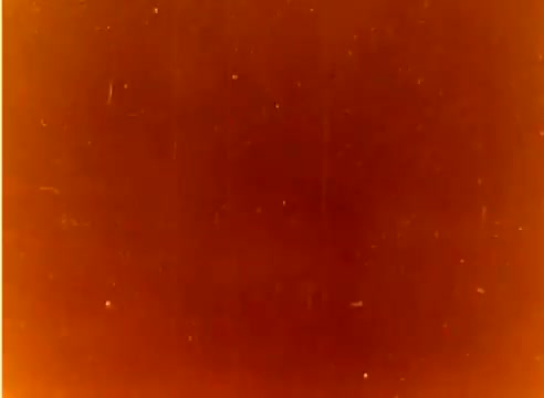 The still appears to be a damaged or degraded frame from a 16mm silent color negative. It has a warm, orange hue with visible scratches and imperfections, indicating its age and the film's wear over time. This frame is part of a sales film highlighting Kodak products leading up to the Christmas season in 1977, featuring Michael Landon and Dick Van Dyke.