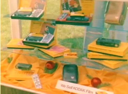 The still from the 'Kodak 1977 Sales Film' showcases a display featuring various Kodak film products. The scene is set with a vibrant yellow cloth underneath, enhancing the presentation of the merchandise, which includes film canisters and promotional materials. The display is neatly arranged, emphasizing the variety of Kodak items available for the upcoming Christmas season. The overall aesthetic reflects the marketing efforts of the time, aimed at promoting Kodak's film offerings.