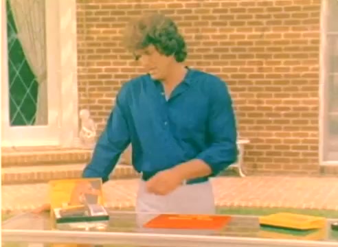 The still features a young man with curly hair, dressed in a blue shirt and light pants, standing beside a table displaying various Kodak film products. The setting appears to be a well-lit outdoor space with a brick wall in the background. This moment likely captures a promotional segment from the 1977 Kodak sales film, showcasing their offerings for the Christmas season. The absence of sound emphasizes the visual presentation of the products.