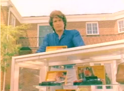 The still features a young man with wavy hair, dressed in a blue shirt, standing in front of a display case filled with colorful products. The background includes a brick building with a balcony, suggesting an outdoor setting. This image is from a silent 16mm color negative used in a Kodak sales film from 1977, promoting products for the upcoming Christmas season.