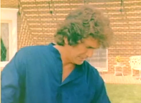 The still captures a moment from the Kodak 1977 Sales Film, showcasing Michael Landon in a casual blue outfit, interacting with a serene outdoor setting. The background features a brick wall and hints of outdoor furniture, reflecting a relaxed atmosphere typical of the era. The image, derived from a silent color negative, emphasizes the vibrant colors and the informal tone intended for promotional purposes leading up to the Christmas season.