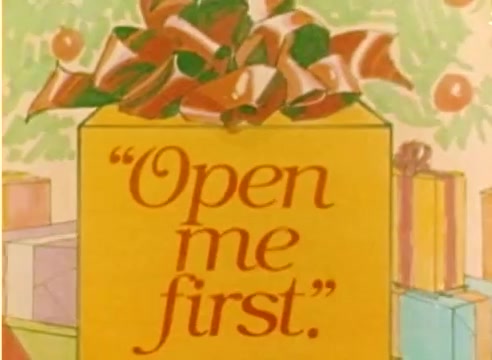 The still from the Kodak 1977 Sales Film features a brightly colored gift box adorned with a large ribbon. The box is labeled 'Open me first.' It is set against a festive background with additional wrapped gifts, emphasizing the holiday theme. The visual plays into the excitement of gift-giving during the Christmas season.