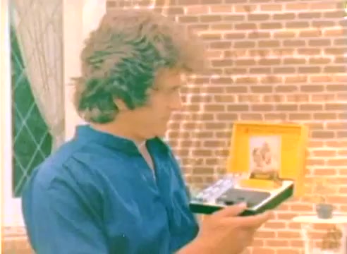 The still captures a moment from the 1977 Kodak sales film featuring a man, likely Michael Landon, displaying a gift box with a vintage camera or photographic equipment. He is wearing a blue shirt and appears engaged with the contents, suggesting a festive or promotional context related to the holiday season. The background features a brick wall, adding to the warm, homey atmosphere typical of Christmas-themed presentations.