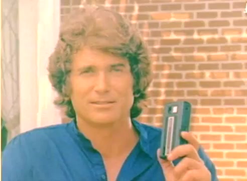 The still captures a young man with wavy hair, dressed in a blue shirt, holding a small device in his hand. The background features a brick wall and light-colored curtains, suggesting an outdoor or well-lit indoor setting. This frame is from a 16mm silent color negative of a Kodak sales film from 1977, likely intended to promote Kodak products during the holiday season. Its vintage aesthetic reflects the era's style.