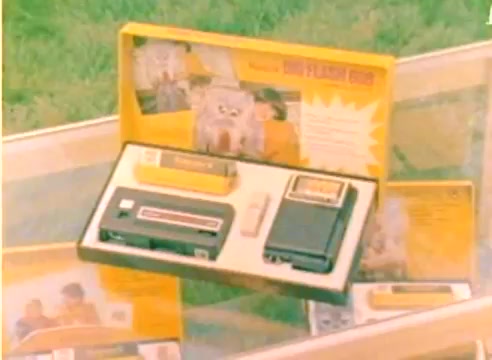 The still depicts a display of Kodak products from the 1977 sales film. Centered is a portable Kodak film projector alongside film reels and promotional materials. The vivid colors and packaging highlight holiday themes, conveying a festive spirit aimed at promoting Kodak's offerings for the Christmas season. The backdrop includes images of families enjoying the products, emphasizing their role in capturing memories.