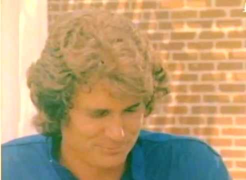The still from the 'Kodak 1977 Sales Film' features a close-up of a man with wavy, medium-length hair, appearing thoughtful or slightly amused. He is wearing a blue shirt and is set against a backdrop that suggests a casual outdoor setting. The lighting is warm, emphasizing a nostalgic ambiance typical of the 1970s. This film was produced as part of promotional content leading into the Christmas season.