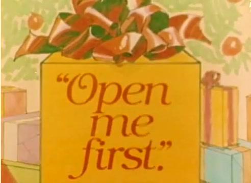 The still features a brightly colored gift box adorned with a large bow, prominently displaying the text 'Open me first.' The background hints at a festive holiday theme, with additional wrapped presents visible, suggesting the context of Christmas and the excitement of gift-giving. The image reflects the spirit of the 1977 holiday season in a whimsical, cheerful manner.