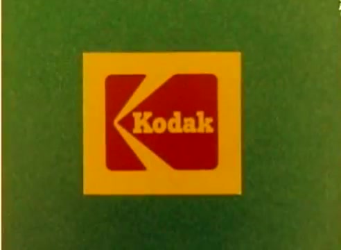 The still features a simple yet striking graphic design showcasing the Kodak logo. The logo is presented in red and yellow on a green background, emphasizing the brand's identity. This image serves as a prominent visual introduction to the 1977 Kodak sales film, likely aimed at promoting their products during the Christmas season. The overall aesthetic is retro and nostalgic, characteristic of the era.