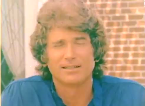 The still features a scene from the 1977 Kodak sales film, showcasing Michael Landon in a relaxed setting. He has medium-length, wavy hair and is wearing a blue shirt. The background includes a brick wall and hints of greenery, evoking a casual outdoor atmosphere. The film is silent and color, likely intended to promote Kodak products during the Christmas season.