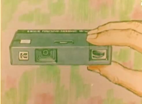 The still depicts a hand holding a Kodak camera, illustrated in a colorful, artistic style. The background features soft, blended colors, enhancing the focus on the camera. This frame is part of a silent sales film from 1977, promoting Kodak products in the lead-up to the Christmas season.