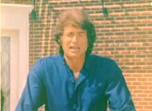 The still from the 'Kodak 1977 Sales Film' features a young Michael Landon wearing a blue shirt, standing in front of a brick wall. The setting appears outdoor, likely in a residential area. His expression conveys enthusiasm or engagement, consistent with promotional content aimed at highlighting Kodak products for the upcoming Christmas season. The film, being a silent color negative, captures the vibrant tones typical of this era's visual style.