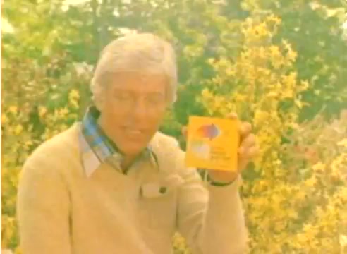 The still depicts a man, likely Dick Van Dyke, in a light-colored sweater holding a box of Kodak film against a backdrop of vibrant yellow flowers. The image is warm-toned, reflecting a nostalgic aesthetic typical of the late 1970s, and captures the promotional spirit of a Kodak sales film aimed at the Christmas season. The absence of sound reinforces the focus on visuals and product presentation.
