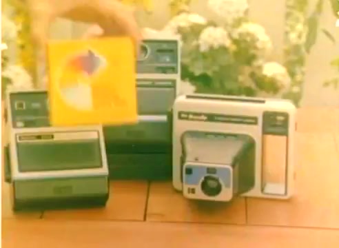The still captures a hand holding a colorful package in front of various Kodak products on a wooden table. These products, likely camera and playback devices, are arranged next to each other, with a backdrop of green plants and white flowers, suggesting an outdoor setting. The overall presentation aligns with the promotional themes of the Kodak 1977 sales film, capturing a festive, inviting atmosphere aimed at the Christmas season.
