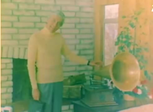 The still from the 'Kodak 1977 Sales Film' features a man in a light sweater standing next to a vintage gramophone. The setting appears cozy, with a brick fireplace and large windows letting in natural light. It evokes a nostalgic holiday atmosphere, promoting Kodak's film during the Christmas season. The wash of color typical of a 16mm silent negative adds a warm, retro quality to the scene.