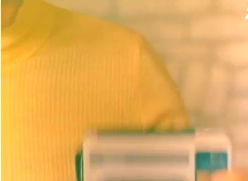 The still features a character wearing a bright yellow sweater, holding a camera. The setting has a warm, soft color palette with a textured background, possibly stone or brick. This 16mm silent negative is part of a Kodak sales film promoting products for the 1977 Christmas season, featuring themes of home and nostalgia.