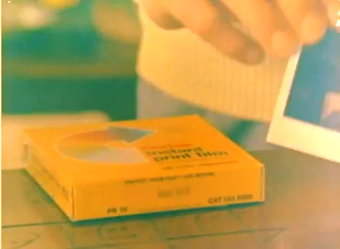 The still features a hand reaching for a photograph near an orange box labeled 'Kodak Color Print Film,' likely showcasing the product. The background is soft-focused, suggesting a cozy, festive atmosphere appropriate for the 1977 Christmas season. The scene conveys a sense of nostalgia associated with film photography.