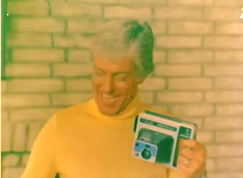 The still shows a man with light-colored hair, wearing a bright yellow sweater and smiling as he holds up a Kodak camera box. The backdrop features a brick wall, and the overall color palette is warm, with a slightly faded appearance indicative of its age as a 16mm silent negative. This image captures a promotional moment likely aimed at highlighting Kodak's products in the lead-up to the 1977 Christmas season.
