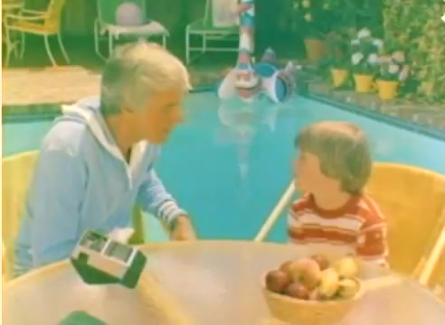 The still features two characters, an adult and a child, sitting at a table near a swimming pool. The adult is wearing a light blue sweater and appears to be engaged in conversation with the child, who is wearing a red and white striped shirt. A basket of fruit sits on the table. The setting is vibrant and colorful, capturing a casual, sunny atmosphere typical of the 1977 era. The scene conveys a warm, familial moment.