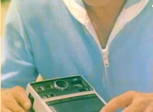The still from the 'Kodak 1977 Sales Film' features a close-up of a man in a light blue zip-up shirt, holding a vintage recording device. His expression is focused on the device, indicative of a presentation or demonstration. The overall scene is brightly colored, typical of the era, capturing a moment likely meant to showcase Kodak's technology. The context suggests it aims to promote their products leading up to the Christmas season.