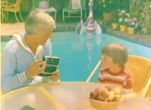 The still features two characters by a poolside table. One, wearing a light blue jacket, holds a camera, while the other, a young boy in a red and white striped shirt, looks on intently. The setting is bright and cheerful, surrounded by greenery and colorful flower pots, creating a warm, inviting atmosphere, typical of mid-1970s family leisure scenes. This moment captures the essence of the Kodak sales film, showcasing family interaction and the joy of photography.