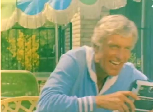 The still from the 'Kodak 1977 Sales Film' features a smiling Dick Van Dyke holding a camera, likely demonstrating it in a casual outdoor setting. He is wearing a light blue sweater, and the background suggests a sunny, festive atmosphere, characteristic of the approaching Christmas season. The scene captures the jovial spirit of the promotional film, aimed to highlight Kodak’s offerings during the holidays.