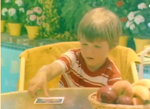 The still features a young boy sitting at a table next to a pool, dressed in a red and white striped shirt. He appears focused as he reaches for a photograph on the table. In the foreground, a basket of apples complements the scene, while vibrant flowers in yellow pots create a colorful backdrop. This captures the lighthearted atmosphere of a Kodak sales film from 1977, designed to evoke a festive mood leading up to Christmas.
