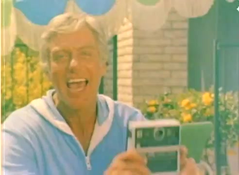The still features a cheerful man in a light blue sweatshirt, holding a camera and smiling broadly. He is likely engaged in a playful moment, possibly promoting Kodak products for the 1977 Christmas season. The background includes bright yellow flowers and a striped awning, contributing to a vibrant, cheerful atmosphere typical of Kodak's family-focused marketing. The image reflects the sunny, nostalgic feel of the era captured in a 16mm silent color negative.