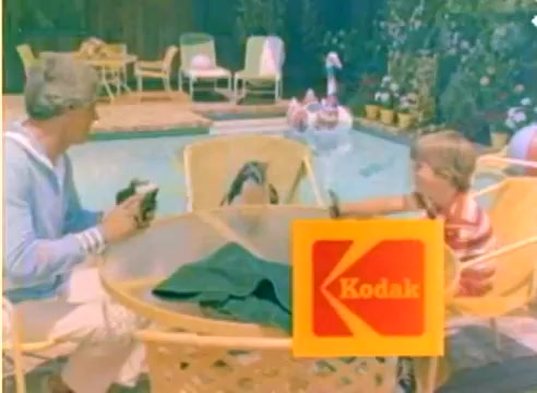 The still features Michael Landon and a young boy seated at a round table beside a swimming pool, enjoying snacks or drinks. Brightly colored lounge chairs are arranged around the pool area, creating a festive, relaxed atmosphere. The scene includes lush greenery and a vibrant Kodak logo prominently displayed in the foreground, indicating a promotional theme for the 1977 Christmas season. The overall tone is cheerful and inviting, typical of family-oriented filmmaking from that era.