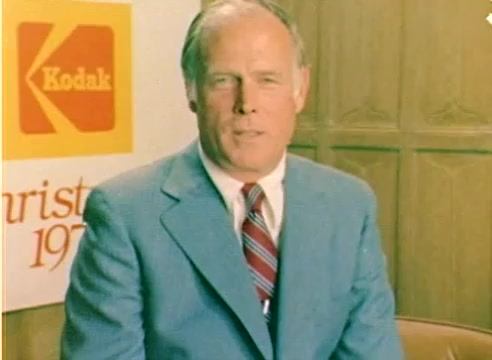 The still features a well-dressed man in a light blue suit seated against a backdrop showcasing Kodak branding and the text 'Christmas 1977.' The setting suggests a promotional context, likely intended for a sales pitch related to Kodak products during the holiday season. The warm color tones and relaxed posture convey a friendly, approachable atmosphere typical of promotional films from that era.