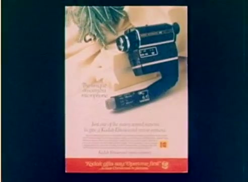 The still from the 'Kodak 1977 Sales Film' features an advertisement for a Kodak video camera positioned against a soft, neutral background. The camera is prominently displayed, showcasing its design and features. The text emphasizes the camera's capabilities and encourages purchasing it as a Christmas gift. The overall aesthetic reflects the promotional style of the late 1970s, combining a festive atmosphere with a focus on family-oriented video recording.
