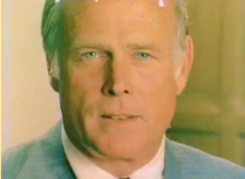 The still is a close-up shot from a 16mm silent negative, showcasing a male figure with light hair and blue eyes dressed in a suit. The background is blurred, suggesting an indoor setting. The color tonality is soft, reflecting the nostalgic feel of the 1977 Kodak sales film context. The image likely emphasizes the warmth and connection associated with the Christmas season.