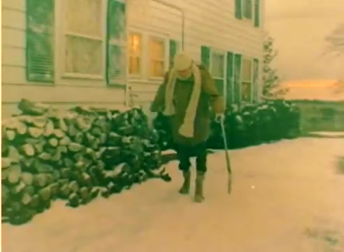 The still from the 1977 Kodak sales film features a snowy outdoor scene. A man, dressed warmly with a scarf and hat, is walking with a cane along a path beside a stack of firewood next to a white house. The atmosphere suggests a serene winter setting, likely intended to evoke a cozy, festive mood for the upcoming Christmas season. The image captures the essence of nostalgia and warmth characteristic of holiday imagery.