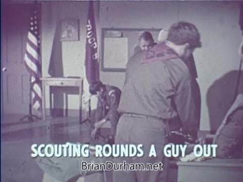 The still from the 16mm PSA 'Scouting Rounds a Guy Out' depicts a scene likely set in a scouting meeting room. It features multiple Boy Scouts engaged in various activities, with an emphasis on teamwork and camaraderie. The background includes American flags and scouting banners, reinforcing the theme of youth development through scouting. The caption at the bottom highlights the focus on personal growth and character building associated with scouting.