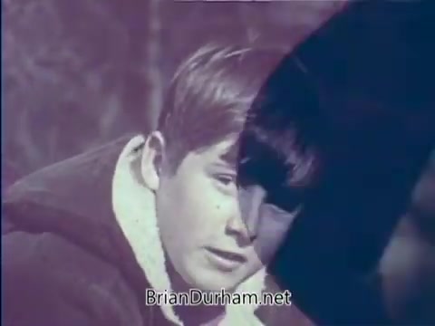 The still from the 'Boy Scout PSA - 'Scouting rounds a guy out'' features a young boy with short hair, wearing a light-colored jacket, seated in a natural setting. He appears thoughtful or introspective. The image has a vintage quality typical of 16mm film, with a subdued color palette. The scene likely emphasizes themes of growth and character development associated with scouting.