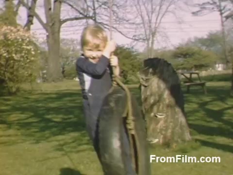 The 16mm still from 'The Tulip Film' captures a cheerful moment in a garden setting, showcasing a child playing with a tire swing. The scene is vibrant, enhanced by beautiful post-war Kodachrome colors, indicative of the era. The background features lush greenery, hinting at the outdoor activities typical of that time. This nostalgic footage predates the famed Tulip Time Festival in Holland, Michigan, making it a valuable piece for flower enthusiasts and those interested in historical home movies.