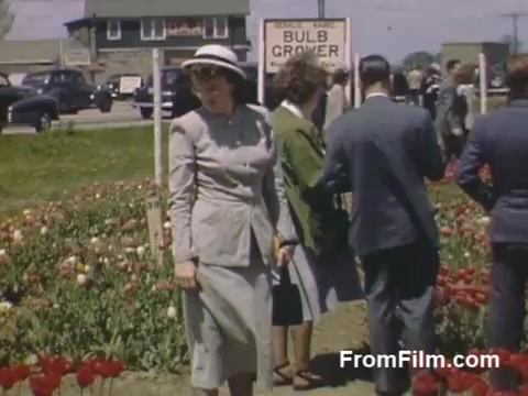 This 16mm still from 'The Tulip Film,' featuring home movies from 1948-1950, showcases vibrant post-war Kodachrome footage of a flower field in Holland, Michigan, before the Tulip Time Festival. The image captures visitors dressed in stylish attire, exploring a blooming garden of tulips, with a sign for a bulb grower in the background. The scene reflects a joyful connection to nature and community amidst the colorful blossoms.