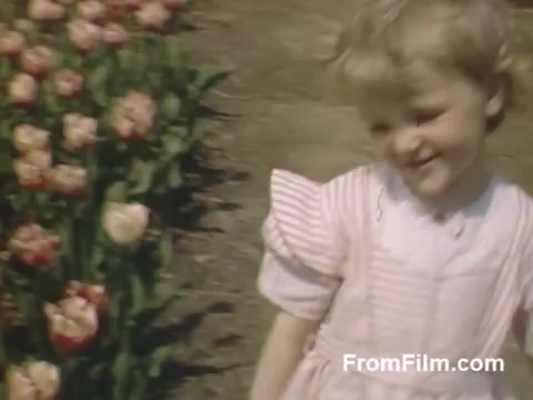 This 16mm still from 'The Tulip Film' features charming post-war Kodachrome footage capturing a young girl in a pink dress, smiling playfully amidst a vibrant display of tulips. The scene is set in Holland, Michigan, highlighting the area's floral beauty, even before the establishment of the renowned Tulip Time Festival. The fresh colors and joyful expression evoke a sense of nostalgia and appreciation for nature.