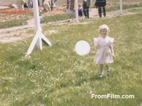 The still from 'The Tulip Film' captures a charming moment from home movies taken between 1948 and 1950. It features a young girl in a light dress happily holding a balloon, set against a backdrop of vibrant tulip fields in Holland, Michigan. The lush green grass and the colorful flowers hint at the beauty of the pre-festival era, showcasing the idyllic atmosphere before the famous Tulip Time Festival began. The post-war Kodachrome quality enhances the nostalgic and vibrant feel of the scene.