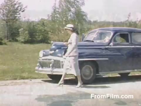 This 16mm still from 'The Tulip Film' presents a snapshot of life in Holland, Michigan during the late 1940s, prior to the establishment of the Tulip Time Festival. It features vibrant post-war Kodachrome footage, showcasing a woman elegantly dressed, stepping out from a classic blue car against a backdrop of lush greenery. The scene captures the charm of the era and the blooming floral landscape, making it a delightful glimpse for flower enthusiasts.